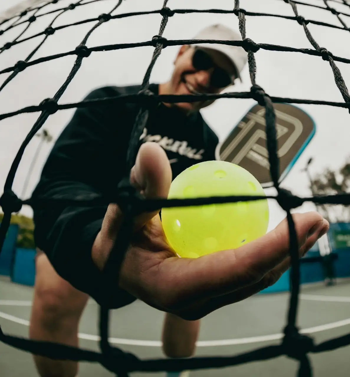 Man holding a pickleball behind a net while smiling