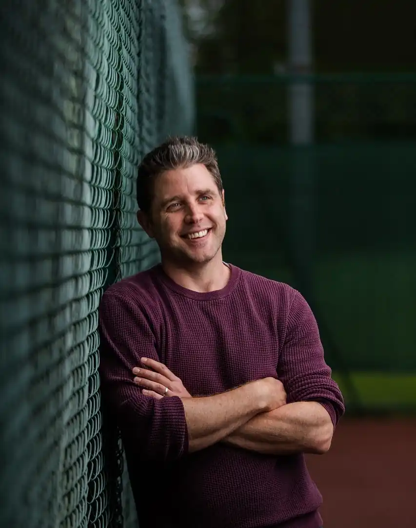 Coach Chris leaning by a tennis court fence in Philadelphia, PA, specializing in adults and competitive improvement.