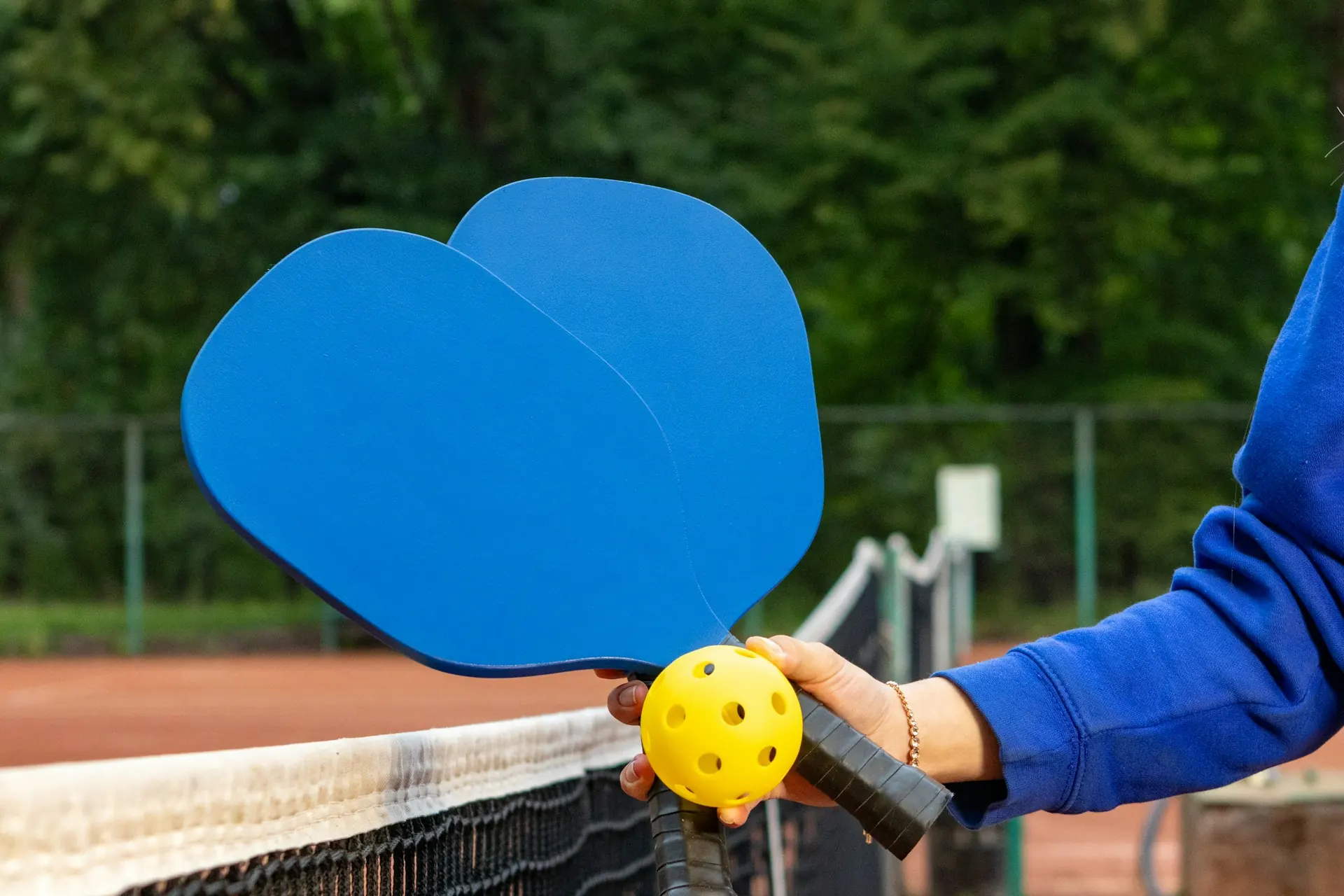 Woman holding two pickleball paddles and a ball in front of a net